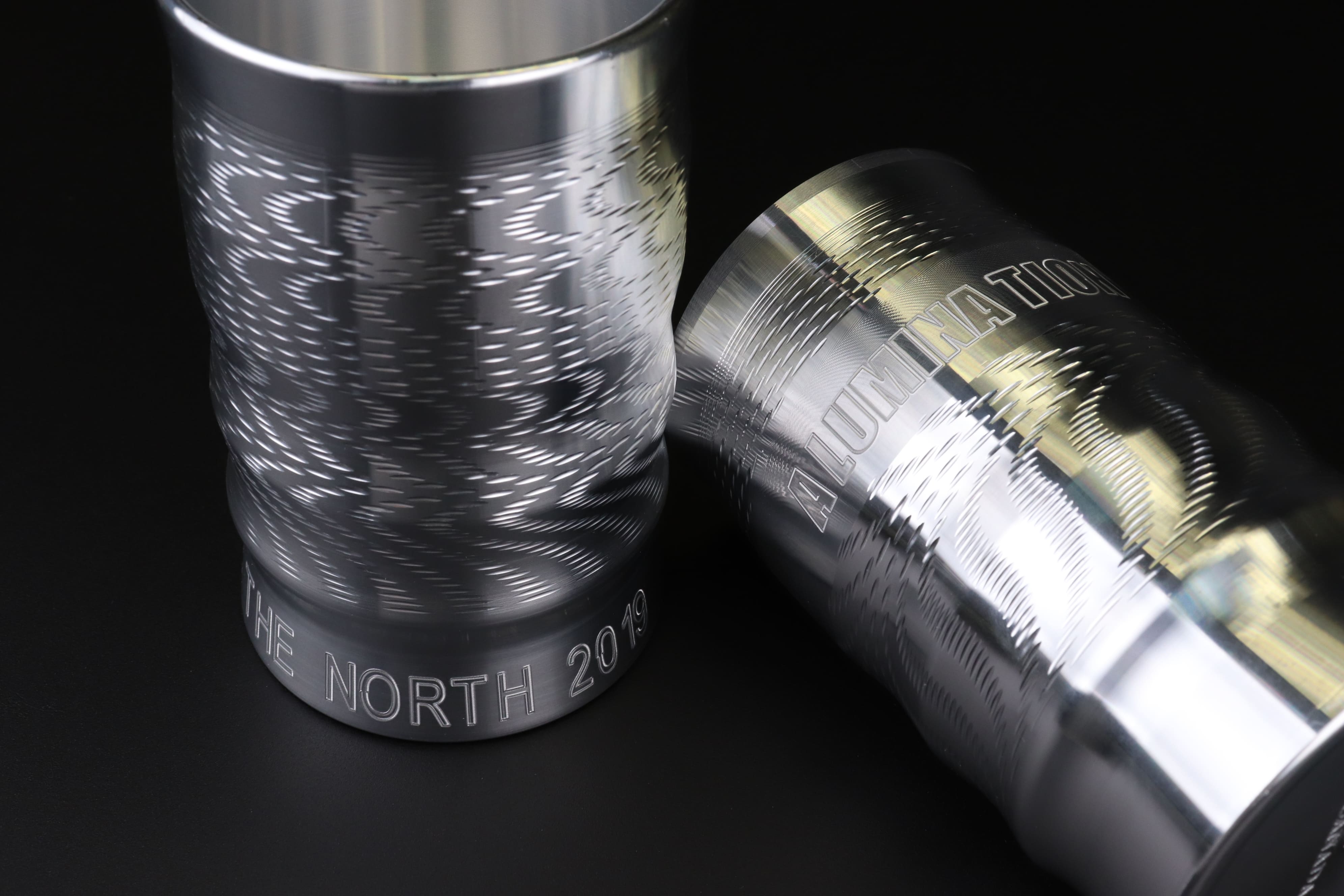 Image of two cylindrical aluminum cups engraved with text 'THE NORTH 2019' for one and 'ALUMINATION' for the other.
