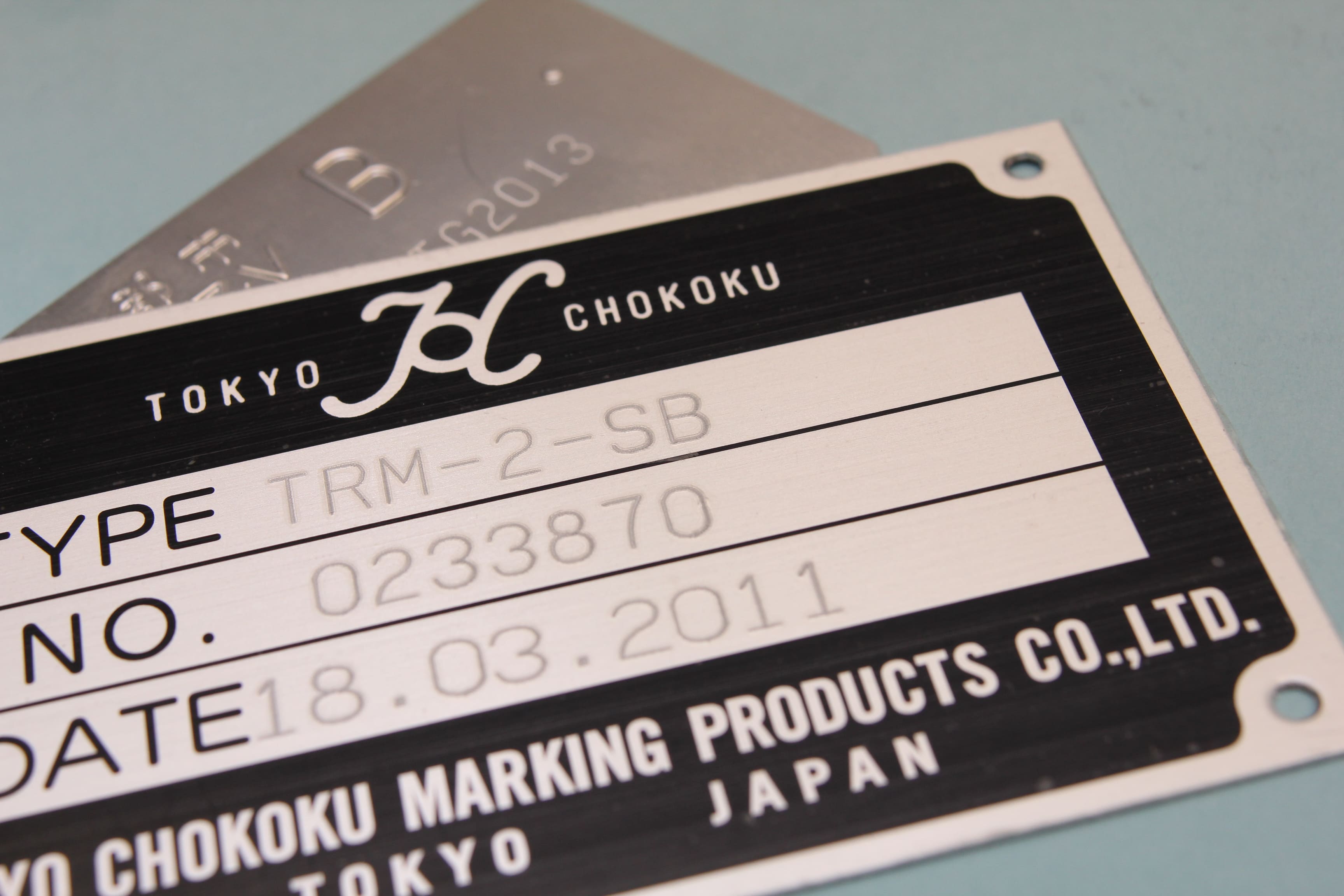 Image of a nameplate engraved with 'TRM-2-SB' for type, '0233870' for number, '18.03.2011' for date, and 'TOKYO CHOKOKU' with logo.