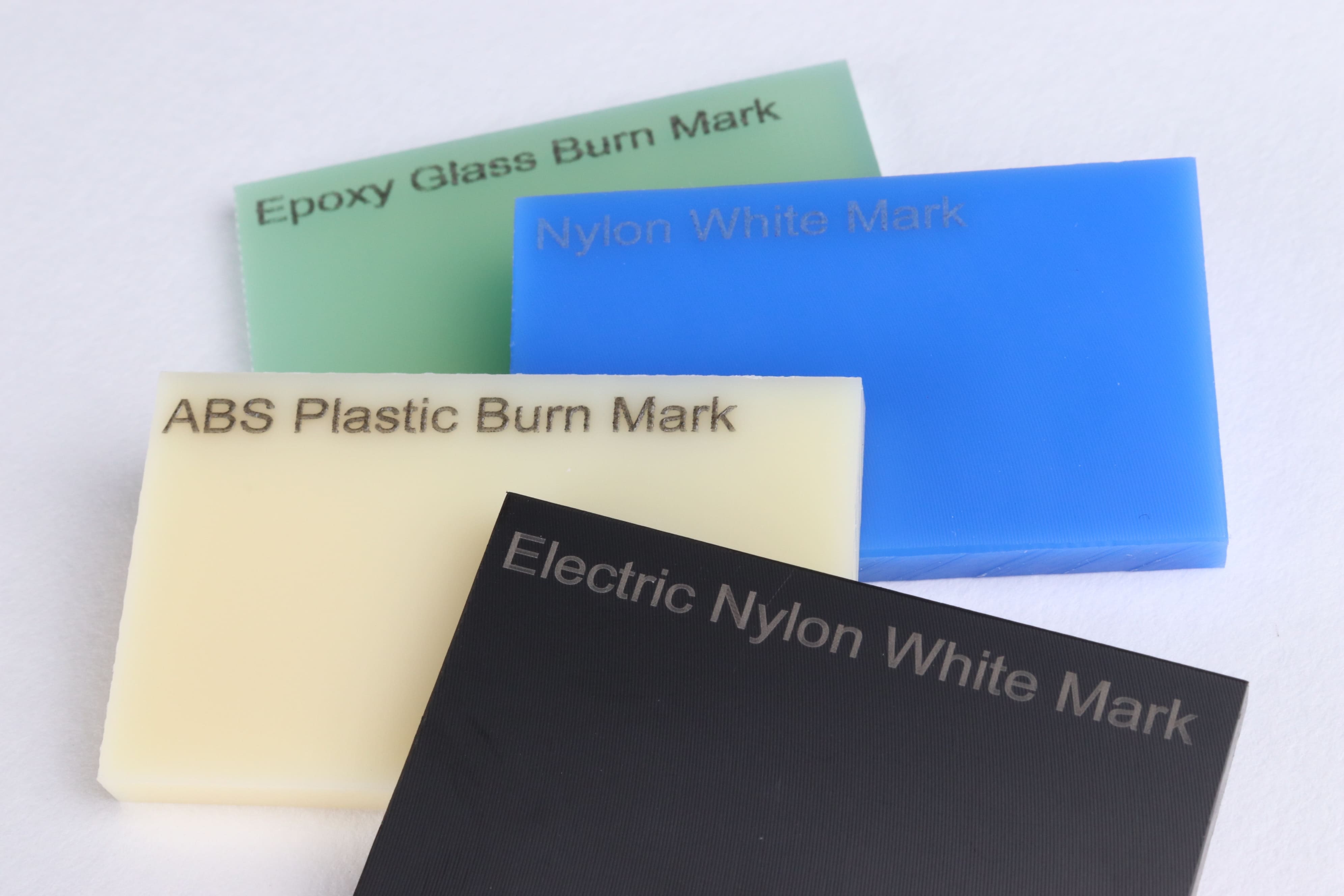 Image of four plastic materials engraved with the text 'Epoxy Glass Burn Mark,' 'Nylon White Mark,' 'ABS Plastic Burn Mark,' and 'Electric Nylon White Mark,' respectively.