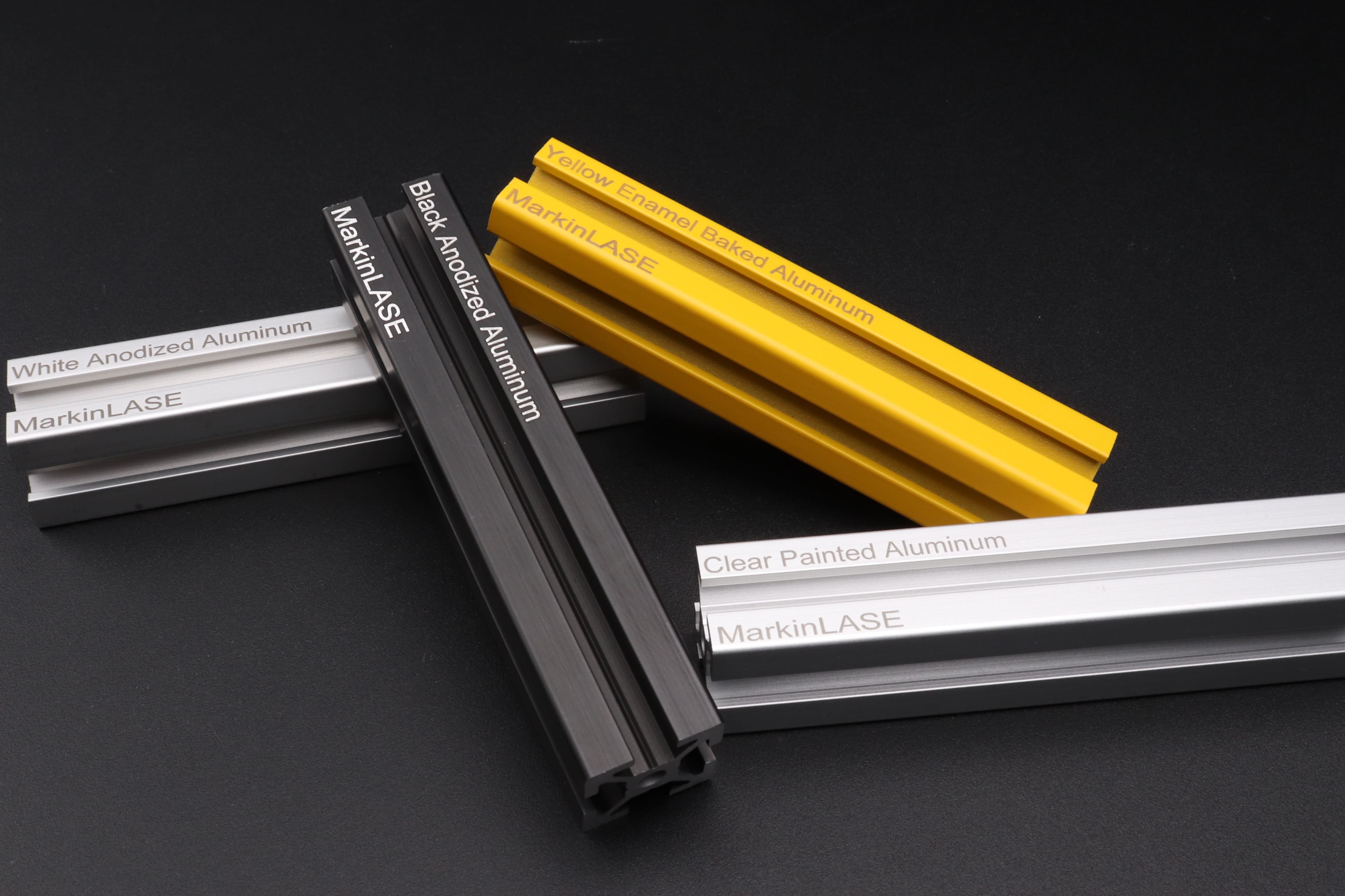 The thin parts of four aluminum materials are engraved with the word 'MarkinLASE' for all and 'White Anodized Aluminum', 'Black Anodized Aluminum', 'Yellow Enamel Baked Aluminum', and 'Clear Painted Aluminum for each'.