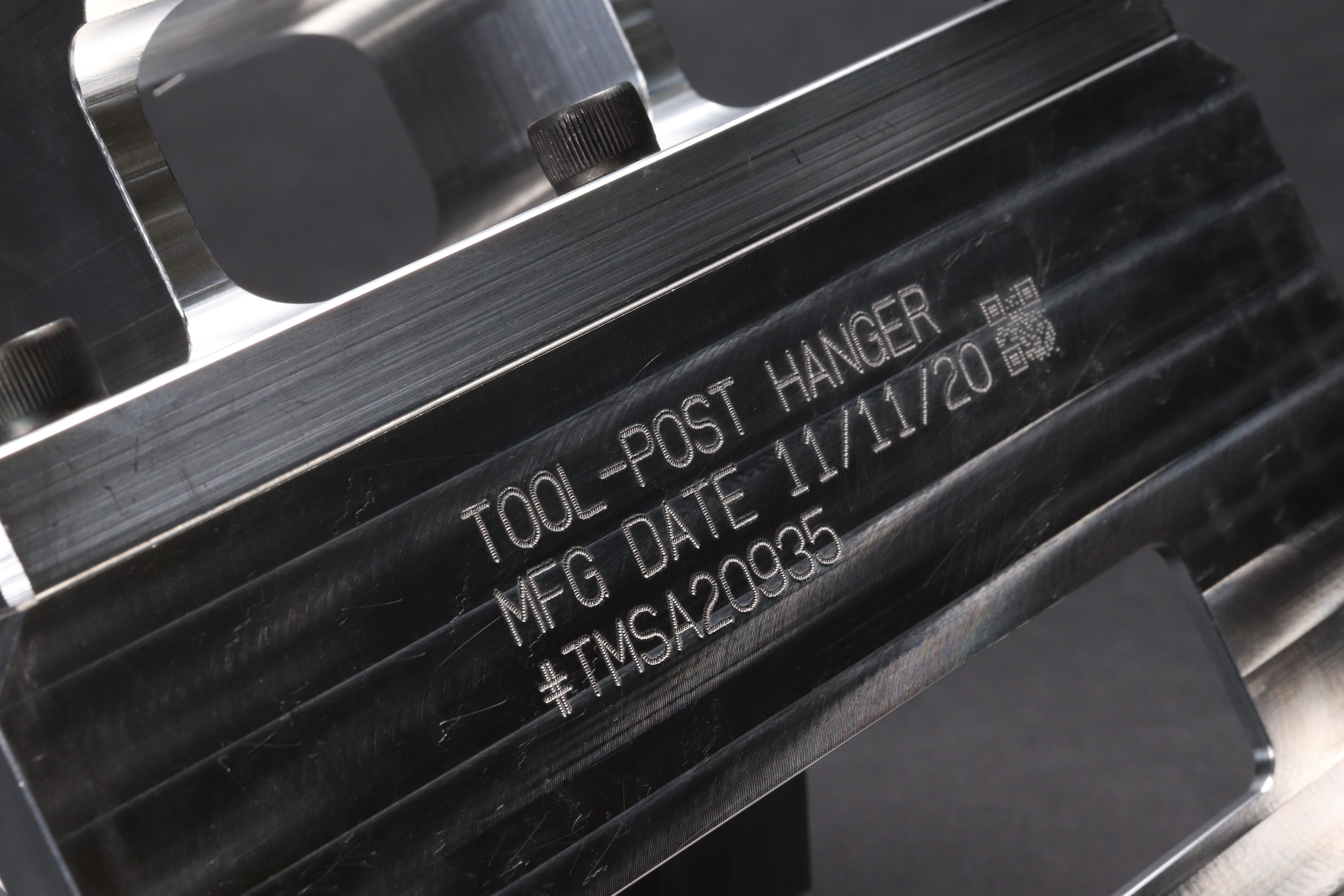 Image of an aluminum fixture engraved with text, numbers and a QR code that reads 'TOOL-POST HANGER MFG DATE 11/11/20 #TMSA20935'.